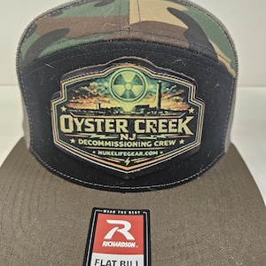 Könnte beinhalten: Eine Flat-Bill-Snapback-Kappe mit Tarnmuster-Oberteil und schwarzem Frontpanel. Die Kappe hat einen Aufnäher mit dem Text "OYSTER CREEK NJ DECOMMISSIONING CREW" und einem Strahlungssymbol. Ein Richardson-Etikett ist angebracht.