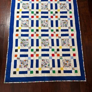 May include: A handmade quilt with a white background, featuring a repeating pattern of blue rectangles and squares. The squares are interspersed with colorful blocks and fabric squares with a road and car design. The quilt has a blue border.
