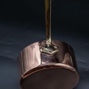 Peut inclure: Une casserole en cuivre poli avec une longue poignée verticale en laiton fixée par une plaque triangulaire à trois rivets. Le côté de la casserole présente des marquages estampillés dont le texte Made in France, avec une surface métallique.