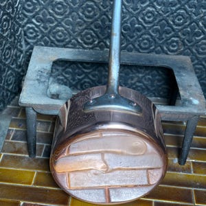 May include: A copper saucepan with a long, silver handle. The pot has a rounded base with a brick-like pattern. The handle is attached with rivets. The pan sits on a small, dark metal stove.