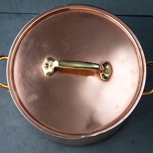 Puede incluir: Una olla de cobre con tapa a juego. La olla tiene un acabado de cobre brillante con asas doradas y un asa dorada en la tapa. La olla es redonda y está sobre una superficie oscura.