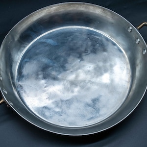 May include: A round, shallow metal pan with a polished silver-toned interior surface and two curved, bronze-colored metal handles attached to the sides with rivets. The pan features a wide, flat base and low, straight walls, presenting a clean, metallic finish.