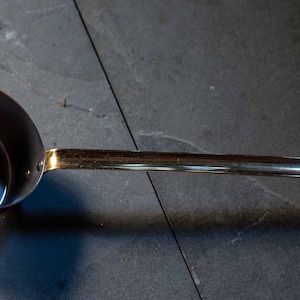 May include: A copper-colored ladle with a long, gold-toned handle. The ladle's bowl is round and deep, with a shiny interior. The handle curves slightly at the end, creating a hook shape. The kitchen utensil is set against a dark background.