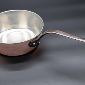 May include: A copper-clad saucepan with a silver interior and a long, riveted handle. The pan has a rounded shape and is designed for cooking. The handle is made of a darker metal, and the pan sits on a dark surface.
