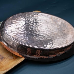 May include: A hammered copper pan with brass handles rests on a wooden tray. The copper pan has an oval shape and a textured surface. The handles are rectangular and attached to the sides of the pan. The wooden tray is light brown.