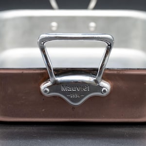 Peut inclure: Un plat à rôtir en cuivre et en acier inoxydable de forme rectangulaire. Le plat a un extérieur en cuivre poli et un intérieur en acier inoxydable. La poignée est en acier inoxydable. Le plat porte l'inscription "Mauviel - 1830".