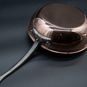 May include: A copper saucepan with a long, riveted iron handle. The pan has a rounded shape and a polished, reflective surface. The handle is dark gray and curves away from the pan's body. The cookware is set against a dark background.