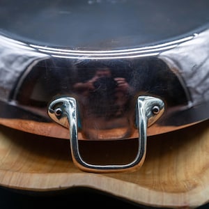 May include: A polished copper pan with a rounded base and a shiny metallic finish. The pan features a sturdy, U-shaped silver-toned metal handle securely attached with two rivets. The exterior surface is reflective, showing a distorted reflection.