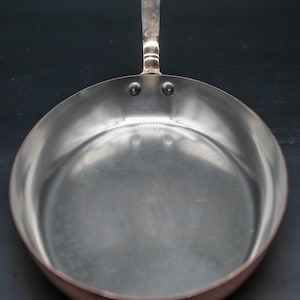 May include: A round copper frying pan with a polished stainless steel interior lining. The pan features a long, flat metal handle attached to the side with two visible rivets. The exterior sides show a warm copper tone with a smooth metallic finish.