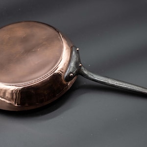 Puede incluir: Una sartén de cobre con un mango largo y oscuro. El interior de la sartén es de un color cobre brillante y reflectante, mientras que el mango parece ser de un material más oscuro. El mango tiene un lazo en el extremo para colgar.