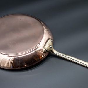 Puede incluir: Una sartén de color cobre con un mango largo plateado. La sartén tiene forma redonda y está hecha de metal brillante. El mango está sujeto con remaches. La sartén está diseñada para cocinar, con un diámetro de aproximadamente 25 cm.