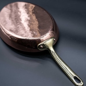 May include: A copper frying pan with a brass handle. The pan has a hammered finish and is oval-shaped. The handle is attached with rivets. The pan is designed for cooking and is a kitchenware item.