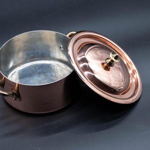 May include: A copper-colored saucepan with a lid, set against a dark background. The lid is slightly ajar, revealing the interior of the pot. The pot has two handles and a shiny, reflective surface.