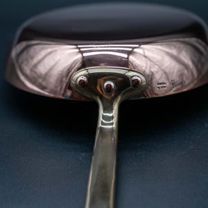 May include: A polished copper frying pan with a smooth, reflective surface and a sturdy metal handle attached by a three-rivet plate. The base of the pan features a small, stamped hallmark near the handle attachment point, showing clear metallic sheen.