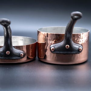 May include: Two copper saucepans with hammered exteriors and dark handles. The pans have a silver interior and are riveted to the copper body. The handles are curved and attached with copper rivets. One pan is smaller than the other.