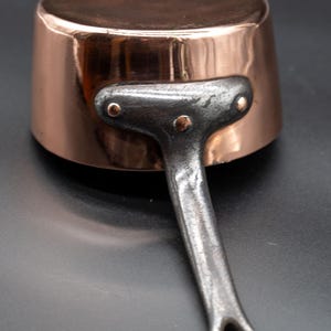 May include: A small copper saucepan with a riveted, cast-iron handle. The pot has a shiny, reflective surface and a dark, matte handle. The handle is attached with two rivets. The pan is designed for cooking.