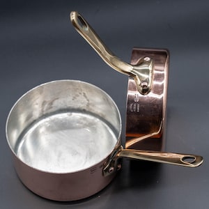 Puede incluir: Dos cacerolas de cobre con asas de latón se muestran sobre un fondo oscuro. Una cacerola está en posición vertical, mostrando su interior, mientras que la otra está de lado. El exterior de cobre contrasta con el interior brillante.