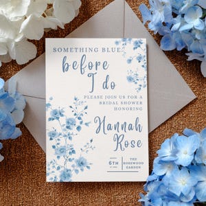 Puede incluir: Una invitación a una despedida de soltera crema con decoraciones florales azules y el texto "Something Blue Before I Do". La invitación incluye los nombres "Hannah Rose" y los detalles del evento. La invitación está sobre un fondo crema texturizado, con un sobre gris.