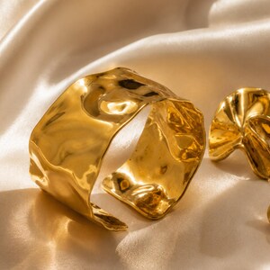 Könnte beinhalten: Ein goldfarbenes Armband mit einer unregelmäßigen, welligen und reflektierenden Oberfläche, begleitet von zwei passenden Ohrsteckern mit einem ähnlichen organischen, gewellten und fließenden Design mit hochglänzender Metalloberfläche.
