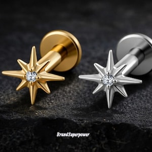 May include: Two star-shaped stud earrings, one gold-toned and one silver-toned, each with a central clear gemstone. The earrings have a flat, circular backing. The gold earring is on the left, and the silver earring is on the right. The background is dark.