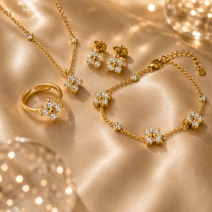 May include: A gold jewellery set consisting of a necklace, a pair of stud earrings, a ring, and a bracelet. Each piece features a four-petaled flower motif encrusted with clear, sparkling stones. The necklace and bracelet include additional small stones.