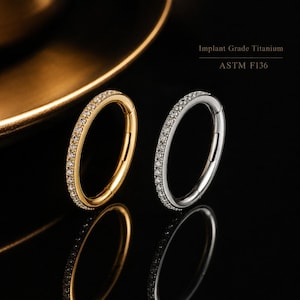 Peut inclure: Deux anneaux circulaires, un doré et un argenté, ornés d'une rangée de petites pierres claires sur le bord extérieur. Le texte Implant Grade Titanium ASTM F136 est visible. Les deux anneaux possèdent un mécanisme de fermeture à charnière.
