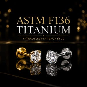 May include: Two threadless flat back studs made of ASTM F136 titanium. One stud is gold-coloured and the other is silver-coloured, both featuring a clear round prong-set gemstone. The text reads ASTM F136 TITANIUM THREADLESS FLAT BACK STUD.