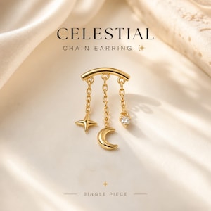 Puede incluir: Un pendiente de cadena dorado con abalorios celestiales. El pendiente presenta una barra curva con tres cadenas colgantes, cada una con un abalorio: una estrella, una luna creciente y una gema en forma de corazón. La palabra "CELESTIAL" está impresa encima.
