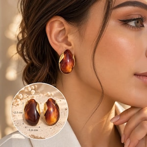 Puede incluir: Un par de pendientes ovalados con una piedra de resina pulida de color ámbar intenso, engastada en un marco de metal dorado liso. Los pendientes miden 2,5 cm de alto y 1,6 cm de ancho, con un acabado brillante y una forma orgánica.