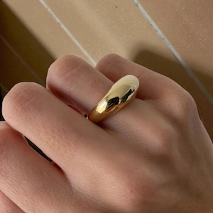 Könnte beinhalten: Ein goldfarbener Ring mit einem glatten, abgerundeten Design. Der Ring hat eine polierte, reflektierende Oberfläche, die das Licht einfängt. Ein schlichtes, aber elegantes Schmuckstück.
