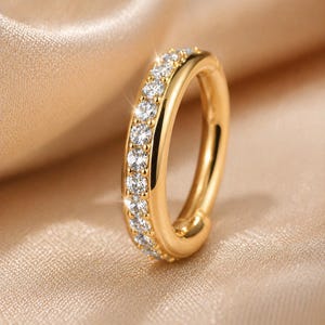Könnte beinhalten: Ein goldfarbener Ring mit einer Reihe klarer, funkelnder Edelsteine, die entlang des äußeren Bandes gesetzt sind. Der Ring wird auf einem weichen, goldfarbenen Stoff präsentiert, was eine luxuriöse und elegante Präsentation ergibt. Das Design des Rings ist schlicht, aber auffällig.