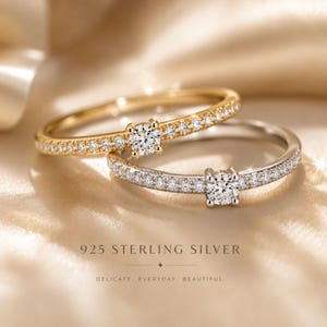 Peut inclure: Deux bagues, une dorée et une argentée, sont présentées. La bague dorée présente un diamant central de taille carrée et de petits diamants le long du sertissage. La bague argentée a un design similaire. Le texte "925 STERLING SILVER" et "DELICATE. EVERYDAY. BEAUTIFUL." sont visibles.
