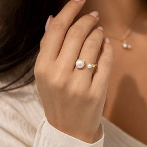 May include: A gold ring with two pearls and small accent stones. The ring has an open design, with a large pearl on one end and a smaller pearl with accent stones on the other. A matching pearl necklace is also visible.