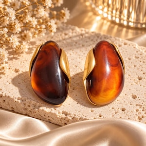 May include: A pair of earrings featuring smooth, polished, oval-shaped amber-colored resin stones. Each stone is encased in a curved, gold-toned metal setting that wraps around the sides, creating a sleek, organic look with warm brown tones.