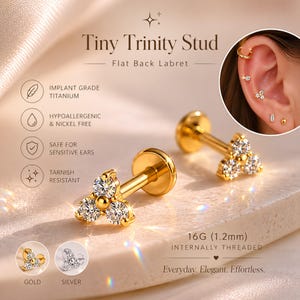 Peut inclure: Un bijou de piercing labret à dos plat avec un motif trinity doré composé de trois pierres rondes transparentes en triangle. Le bijou est de calibre 16G (1,2 mm) et possède un filetage interne. Le texte Tiny Trinity Stud est visible.