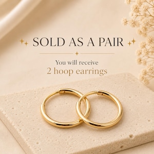 Puede incluir: Un par de pendientes de aro dorados con un acabado liso y pulido. El texto sobre los pendientes dice: SOLD AS A PAIR. You will receive 2 hoop earrings. Los pendientes son circulares con un diseño sencillo y continuo con cierre de bisagra.