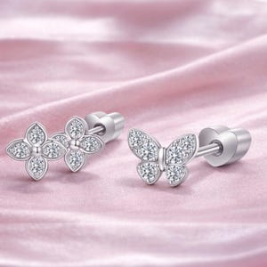 Peut inclure: Deux boucles d'oreilles à clous en argent. L'une présente un motif de fleur à quatre pétales, l'autre une forme de papillon. Les deux sont ornées de petits cristaux étincelants. Le fond est un tissu rose tendre.