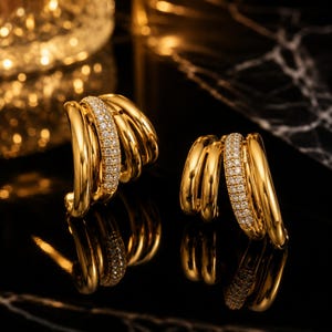 Könnte beinhalten: Ein Paar goldene Creolen mit mehrschichtigem Design. Jeder Ohrring besteht aus drei glatten, polierten Goldreifen, die ein mittleres Band flankieren, welches mit kleinen, klaren, funkelnden Steinen besetzt ist und einen geschichteten Look erzeugt.