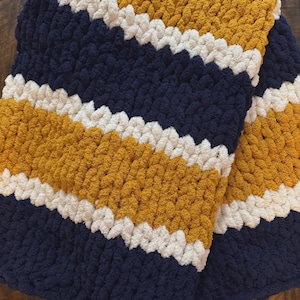 May include: A chunky knit blanket with alternating stripes of navy blue, mustard yellow, and white. The blanket is folded, showcasing the texture and color pattern. The cozy, handmade blanket is perfect for snuggling.