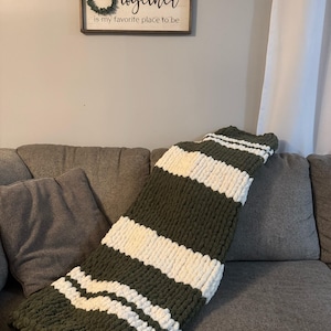 May include: A chunky knit blanket in olive green with wide white stripes, draped over a gray sofa. The blanket's texture is visible, suggesting a cozy and warm feel. A wooden sign with the text "together is my favorite place to be" hangs on the wall.