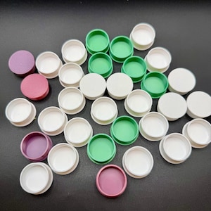 May include: An assortment of small, round, colorful plastic lids and containers. The lids are in shades of white, green, purple, and pink. The containers are white. The items are arranged on a black surface.