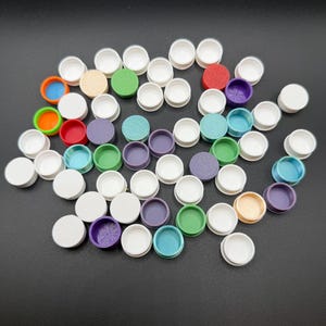 May include: A collection of small, colorful containers with white lids. The containers are in various colors, including red, green, purple, and blue. The lids are white. The containers are scattered on a dark surface.