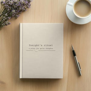May include: A light beige hardback journal with the text "Tonight's ritual" and "a place for quite thoughts" printed in black on the front cover. The journal is positioned next to a silver metal pen, a white coffee cup, and purple flowers.