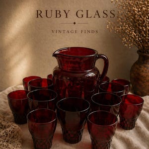 Puede incluir: Un juego de cristalería de color rojo rubí, que incluye una jarra y varios vasos. La jarra tiene un asa y un pico vertedor. Los vasos son cortos y tienen un diseño texturizado. El texto dice "RUBY GLASS VINTAGE FINDS".