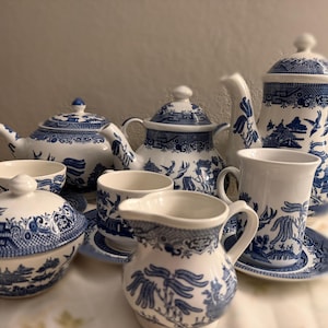 Könnte beinhalten: Ein Set aus blau-weißem Porzellan, darunter Teetassen, Teekannen, ein Sahnekännchen und eine Zuckerdose. Das Willow-Muster zeigt detaillierte Landschaftsszenen. Das Teeservice ist auf einer floralen Tischdecke angeordnet.