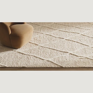May include: Cream-coloured area rug featuring a geometric diamond pattern. The rug has a textured, woven appearance. A section of a tan armchair is visible in the upper left corner, suggesting a living room setting. The rug's design adds a touch of elegance.