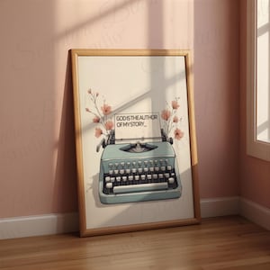 Puede incluir: Un cuadro enmarcado con una máquina de escribir vintage turquesa y el texto "GOD IS THE AUTHOR OF MY STORY". La obra de arte incluye detalles florales y está sobre un fondo rosa claro, con un marco de madera.