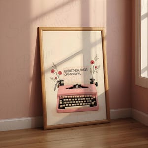 May include: A framed print showing a pink vintage typewriter with the text "GOD IS THE AUTHOR OF MY STORY." The artwork includes floral accents and a wooden frame. It leans against a pink wall and wooden floor.