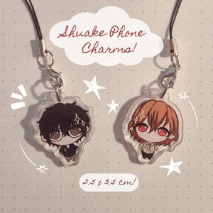 Peut inclure: Deux breloques de téléphone en acrylique transparent avec des illustrations de personnages de style chibi, l'un aux cheveux noirs avec des lunettes, l'autre aux cheveux bruns et yeux rouges. Texte : Shuake Phone Charms! 2.5 x 3.5 cm!