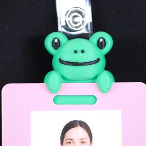 Puede incluir: Un porta-credenciales verde con forma de rana con una sonrisa y ojos negros, sujeto a una tarjeta de identificación rosa. La tarjeta de identificación tiene un clip de plástico transparente y una foto de una persona. El fondo es negro.